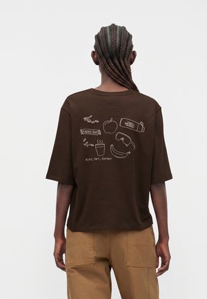 Person med flettet hår bærer en brun t-shirt med hvide illustrationer af æble, banan, energibar, kaffe, goggles og teksten "Hike, Eat, Repeat" på ryggen.