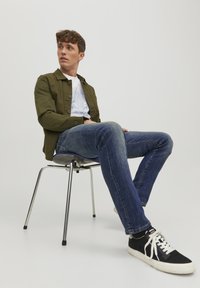 Giacca botton-up verde oliva sopra una t-shirt grafica bianca, jeans blu e sneakers nere con lacci bianchi, seduto su una sedia in cromato.