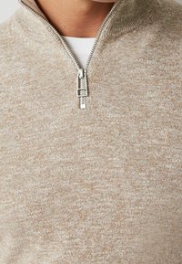 Beżowy sweter na zamek z fakturowanym wzorem dzianiny i wysokim kołnierzem. Srebrny zamek błyskawiczny i subtelny detal z emblematem przy dolnym brzegu.