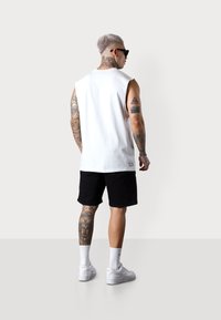 White sleeveless shirt, black shorts, white sneakers, and tattoos visible on arms and legs. Model viewed from the back, wearing sunglasses.