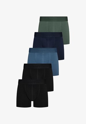 Jonge man staat op blote voeten in groene boxershorts, kijkt naar links met zijn rechterhand op zijn heup, tegen een effen achtergrond.