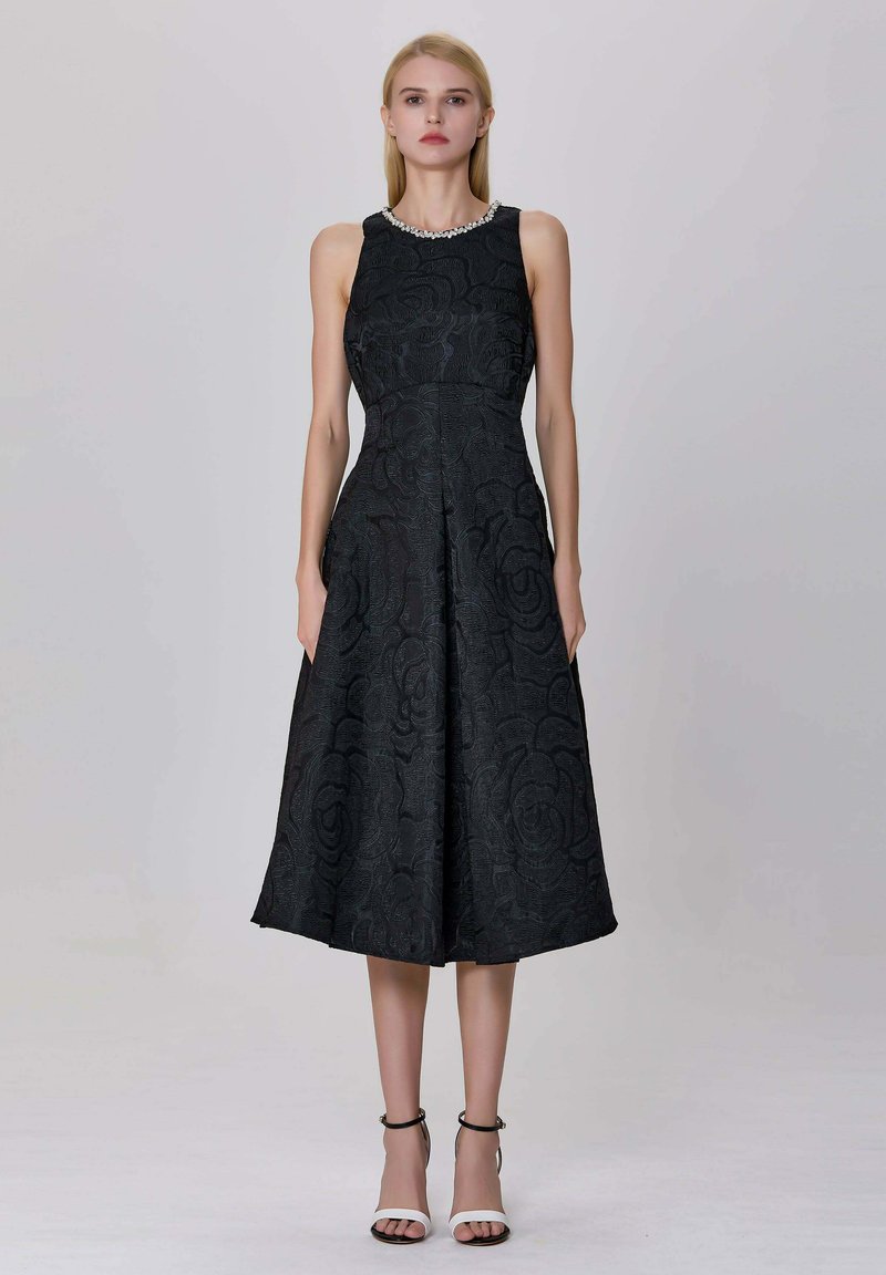 Femme portant une robe midi noire sans manches avec une texture à motif floral et un col orné de perles, accompagnée de sandales à brides de cheville noir et blanc.