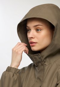 Giacca impermeabile color khaki con cappuccio, caratterizzata da polsini elastici, chiusura con zip e dettagli a pressione. Texture liscia, design elegante.