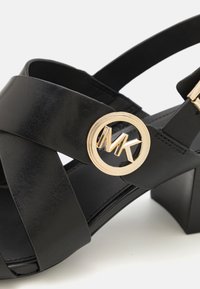 Sandale en cuir noir avec brides croisées, boucle circulaire dorée avec logo MK, et talon bloc sur fond blanc.
