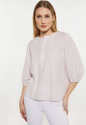 Femme blonde portant un chemisier rayé rose clair et blanc avec des manches bouffantes trois-quarts et un pantalon blanc, debout devant un fond blanc.