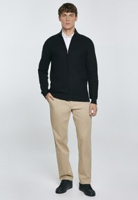 Svart zip-up tröja med långa ärmar, kombinerad med beiga byxor och svarta sneakers. Slät tygkvalitet, figurnära design, minimalistisk stil.