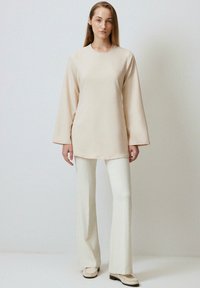 Long-sleeve cream tunic top with a round neckline, paired with white flared pants and light-colored shoes, presenting a minimalist design.