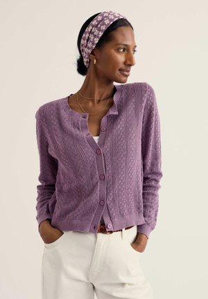 Femeie purtând un cardigan texturat lavandă cu nasturi, pantaloni albi, cercei aurii în formă de cerc, coliere suprapuse și bentiță cu flori, cu mâinile în buzunare, uitându-se spre dreapta.