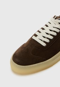 Basket en daim marron avec des lacets blancs et une semelle en caoutchouc translucide, présentant des détails de couture subtils et un design de bout rond.