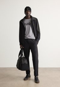 Dark, lightweight jacket with gray detailing, gray tee with "JUG" text, black pants, black sneakers, and a black beanie, holding a bag.