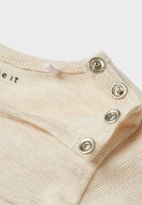 Tissu en coton beige clair avec un tissage texturé. Présente des boutons-pression en argent à l'épaule et une étiquette avec "love it" imprimée à l'intérieur.