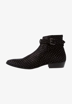 Zwarte suède enkellaars met puntige neus, kleine gouden studs, lage hak en verstelbare band met zilveren gesp.