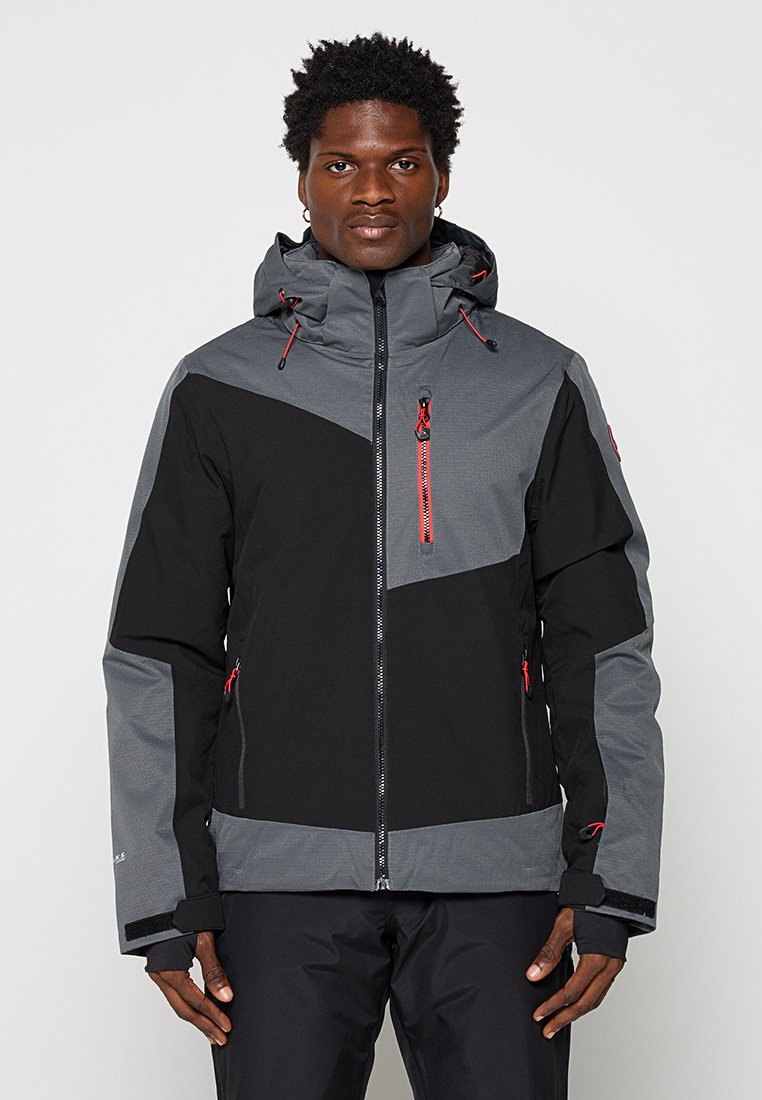 Hombre vestido con una chaqueta negra y gris con detalles en rojo en el cierre, de pie de frente contra un fondo claro y liso.