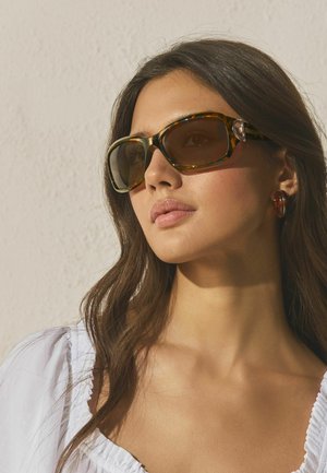 Frau mit langen braunen Haaren, die große Sonnenbrillen mit Schildpattmuster und bernsteinfarbene Creolen trägt, gekleidet in einem weißen Oberteil vor einem schlichten Hintergrund.