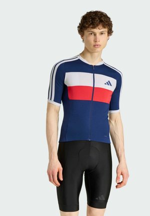 Jeune homme portant un maillot de cyclisme bleu marine, blanc et rouge avec fermeture éclair, ainsi qu'un cuissard de cyclisme noir rembourré, debout devant un fond uni.