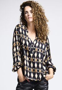 Schwarze Bluse mit geometrischem Gold- und Creme-Muster, V-Ausschnitt, lockeren Ärmeln und glatter Textur. Kombiniert mit dunklen Jeans.