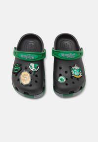Black clogs with green accents, featuring assorted decorative pins. Made from lightweight material, with ventilation holes and a flexible strap.