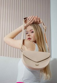 Béžová kožená crossbody taška s prohnutým klopou, zlatými detaily a nastavitelným strapcem, umístěná na pozadí s světle pruhovaným vzorem.