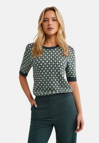 Maglione a maniche corte di colore turchese con un motivo geometrico verde chiaro. Collo e orlo a costine. Abbinato a pantaloni verdi coordinati.