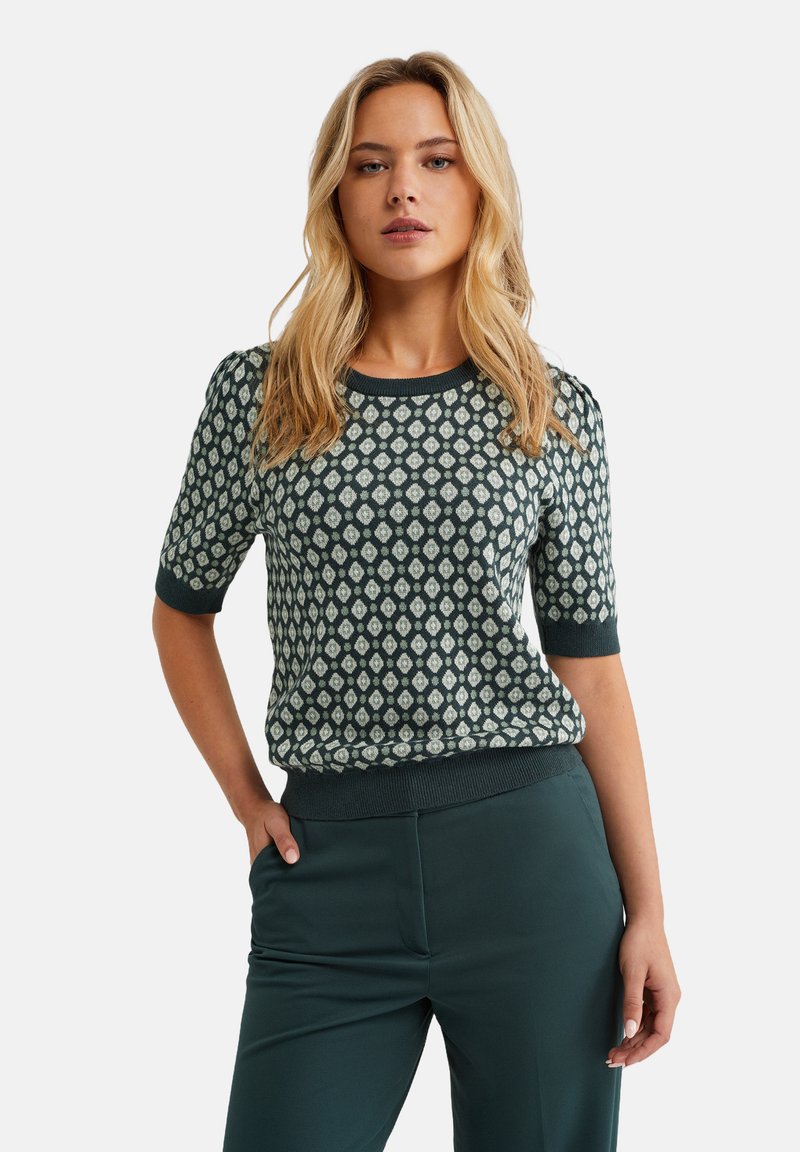 Maglione a maniche corte di colore turchese con un motivo geometrico verde chiaro. Collo e orlo a costine. Abbinato a pantaloni verdi coordinati.