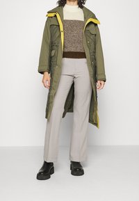 Grøn quiltet parka med gult trim, båret over en brun og beige strikket sweater, parret med lysegrå bukser og sorte platform støvler.