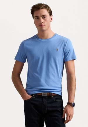 Camiseta de algodón azul con cuello redondo y mangas cortas, que presenta un pequeño logotipo bordado en el lado izquierdo del pecho, combinada con jeans oscuros.