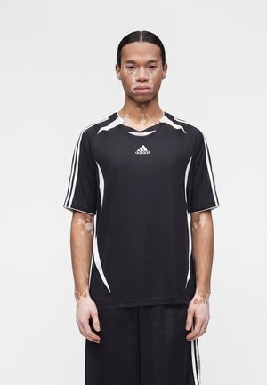 Άνδρας με λεύκη που φοράει μαύρο αθλητικό μπλουζάκι και σορτς Adidas, στέκεται μπροστά σε απλό λευκό φόντο, στραμμένος προς τα εμπρός.