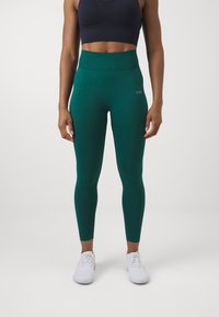 Green leggings made of stretchy material with a ribbed waistband, featuring flatlock seams and a logo on the hip. Paired with white athletic shoes.