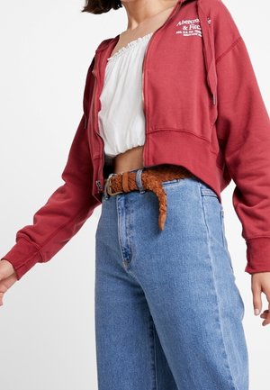Femme portant un sweat à capuche zippé rouge sur un top court blanc, un jean taille haute bleu et une ceinture tissée marron.