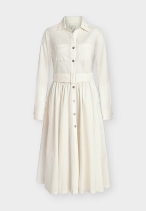 Femme en robe midi ceinturée à manches longues couleur écrue, tenant un sac à main beige, portant des mules à talons ouvertes noires, debout sur fond blanc.