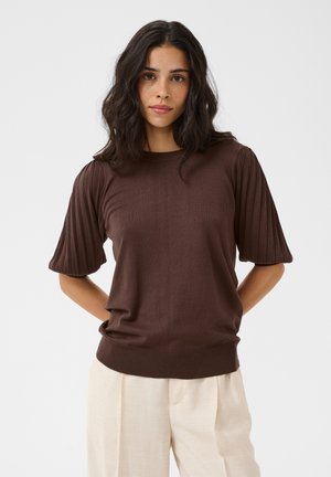Femme aux cheveux bruns ondulés porte un pull côtelé marron à manches courtes et un pantalon ample beige clair, debout les mains derrière le dos.