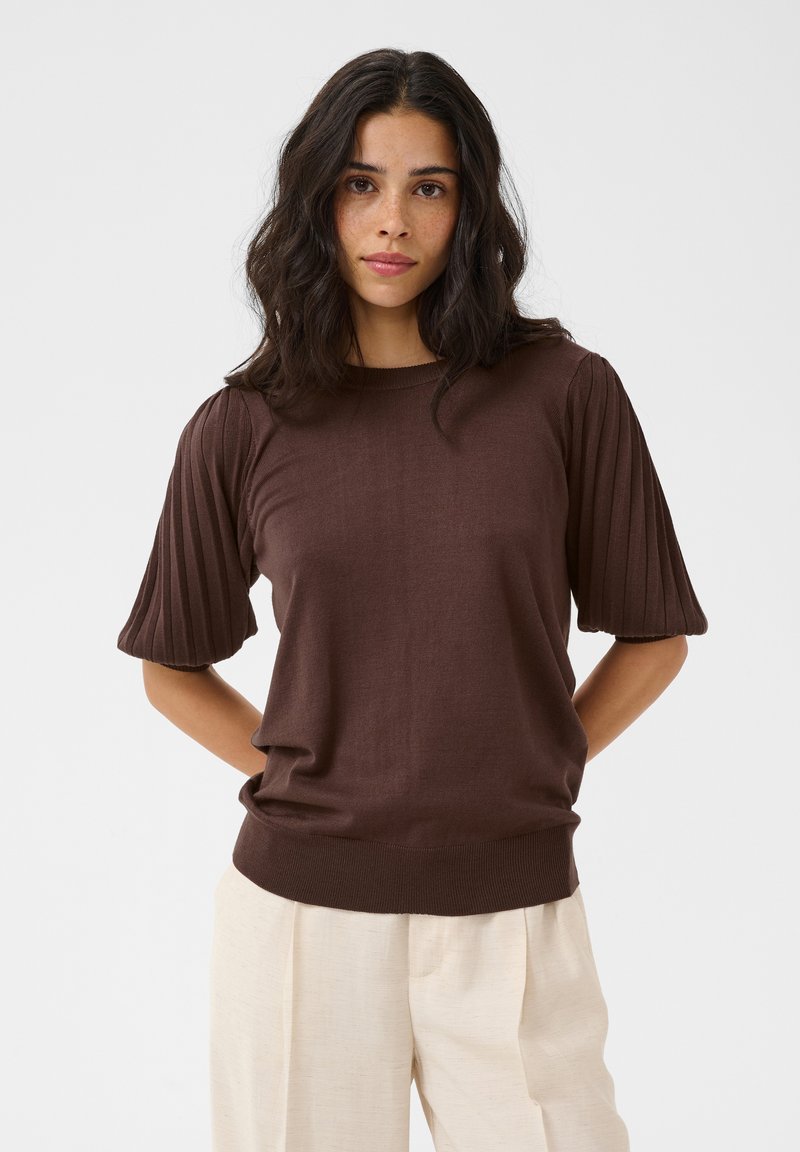 Femme aux cheveux bruns ondulés porte un pull côtelé marron à manches courtes et un pantalon ample beige clair, debout les mains derrière le dos.