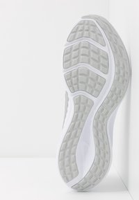 Podešev bílé sportovní obuvi s texturovanou šedou gumovou podrážkou zobrazená vertikálně proti bílému rohovému pozadí.