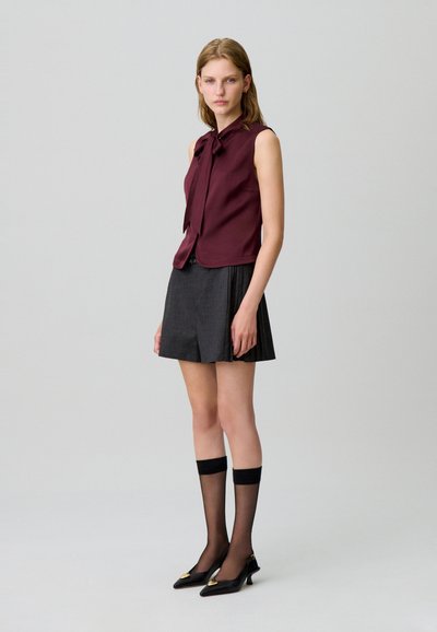 Blusa senza maniche bordeaux con dettaglio fiocco, abbinata a una gonna plissettata nera, calze al ginocchio e décolleté neri a punta con accenti dorati.