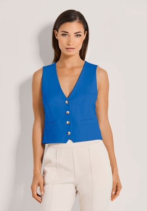 Frau mit glattem braunem Haar, die eine ärmellose königsblaue, geknöpfte Weste und weiße, hoch taillierte Hosen trägt und vor einer hellen Wand steht.