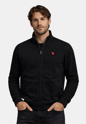 Man wearing black zip-up jacket with red embroidered logo on chest, hands in pockets, neutral background.