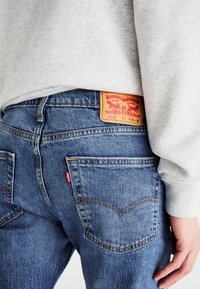 Personne portant un jean denim bleu Levi's 515 avec une étiquette de logo rouge visible et un patch de marque en cuir marron, associé à un sweat-shirt gris clair.
