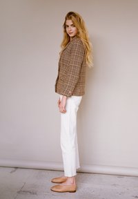 Blazer a quadri marrone con una vestibilità su misura, pantaloni bianchi a 3/4 e scarpe beige con tacco basso. Modella in posa di lato su uno sfondo neutro.