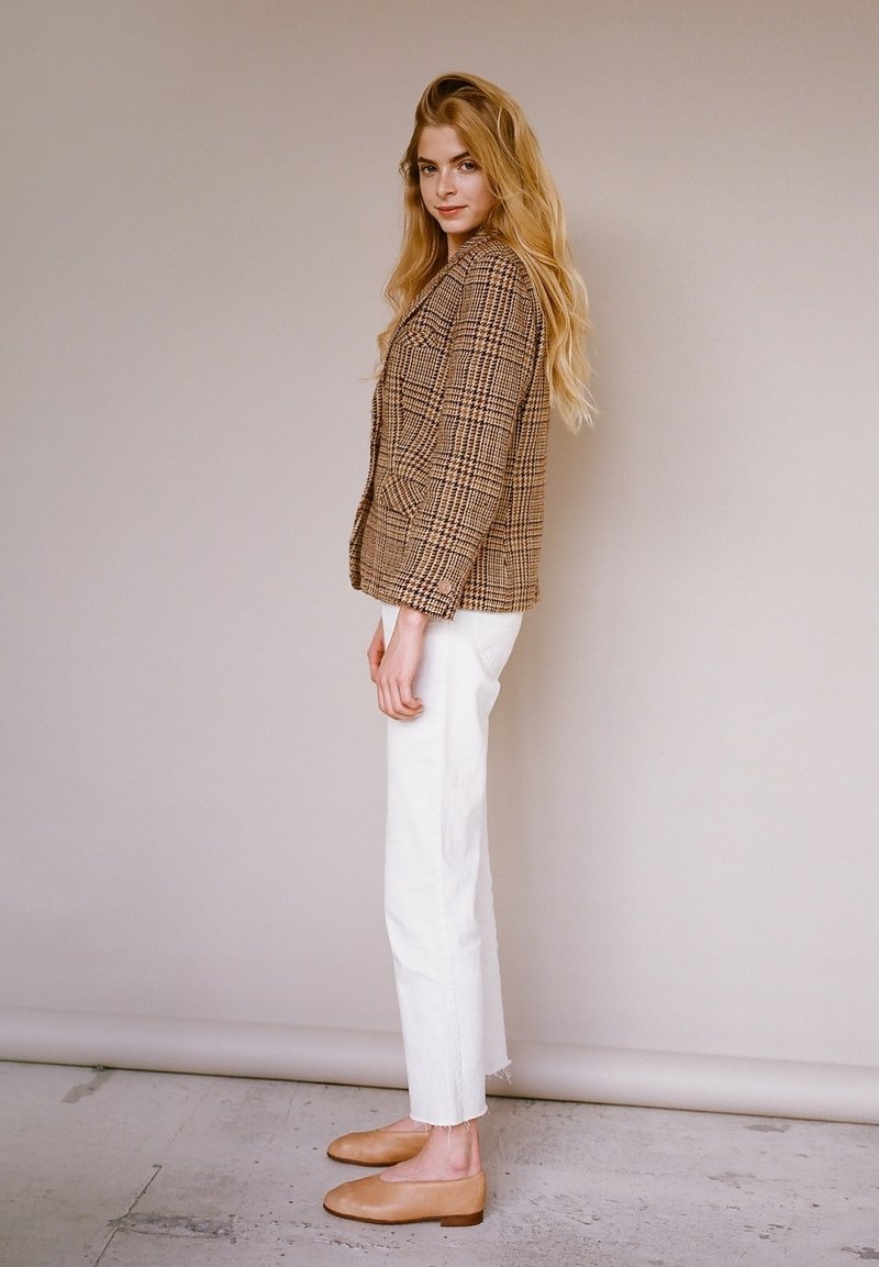 Kvinde med langt blond hår iført en brun ternet blazer, hvide bukser og beige flade sko, stående sidelæns mod en ensfarvet beige væg.