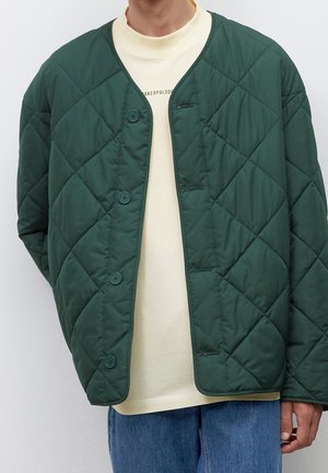 Veste matelassée verte avec un col côtelé, fermeture à boutons et motif en losanges ; portée sur une chemise jaune clair et un jean bleu.