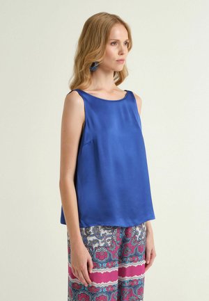 Femme portant un haut bleu roi sans manches et un pantalon ample à motifs rose, bleu et blanc, se tenant avec une expression neutre.