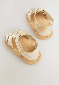 Gold leather infant sandals with a woven design, open back, and cushioned tan footbed. Textured surface with small floral inner pattern.