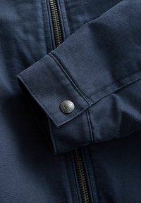 Chaqueta azul marino con una tela texturizada, que presenta una cremallera y un detalle de broche metálico en el puño para el cierre. Primer plano del diseño de la manga.