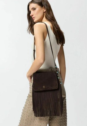 Młoda kobieta w białym prążkowanym topie na ramiączkach i beżowych spodniach z ćwiekami, niosąca ciemnobrązową torebkę na ramię z frędzlami.