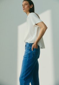 Žena stojaca s rukou v zadnej ponke, oblečená v bielej blúzke s krátkym rukávom a modrých džinsách, na jednoduchom svetlom pozadí.