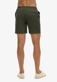 Mann steht und zeigt die Rückseite olivgrüner Shorts mit Reißverschlusstasche, trägt beigefarbene Sneakers vor weißem Hintergrund.