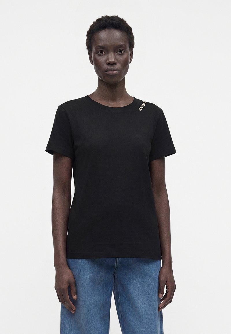 Femme portant un t-shirt noir uni à manches courtes et un jean bleu ample, debout devant un fond blanc.