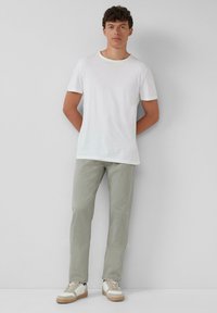 Jonge man die tegen een muur staat in een eenvoudig wit T-shirt, lichtgrijze broek en witte sneakers met rubberen zolen.