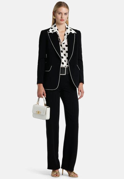 Abito nero su misura con piping bianco, blusa a pois, pantaloni palazzo, sandali con dettagli dorati e una piccola borsa bianca.