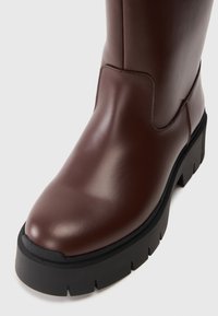 Botín marrón impermeable con un acabado liso, punta redondeada y suela gruesa negra que presenta un patrón texturizado para mayor tracción.